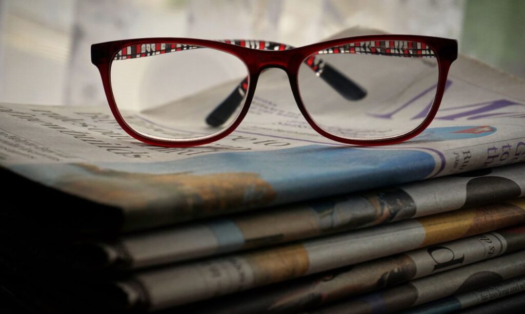 Home Close-up of red glasses resting on stacked newspapers, emphasizing reading and vision.