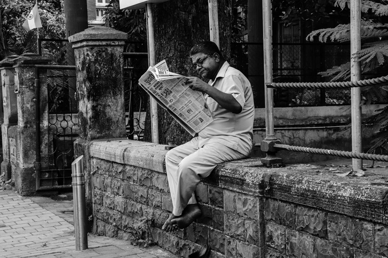 Black and white photo of an elderly man reading a newspaper while sitting on a wall in a street in Mumbai.