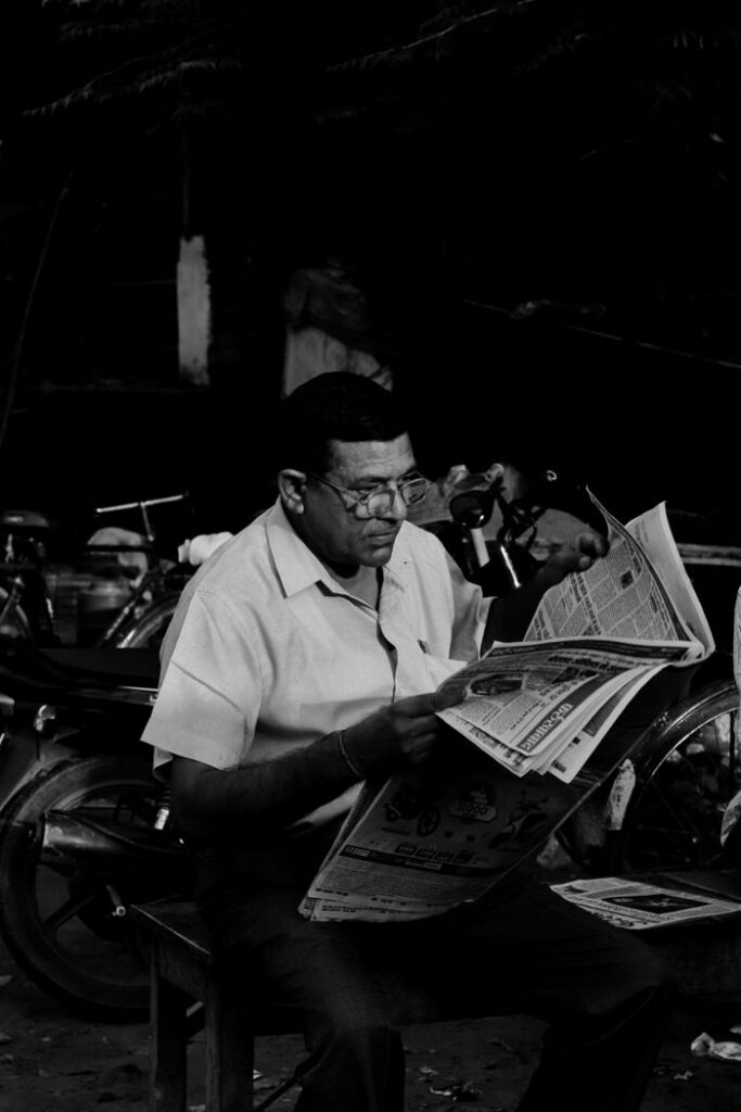 Black and white photo of a man sitting outside reading a newspaper, showcasing leisure and focus.